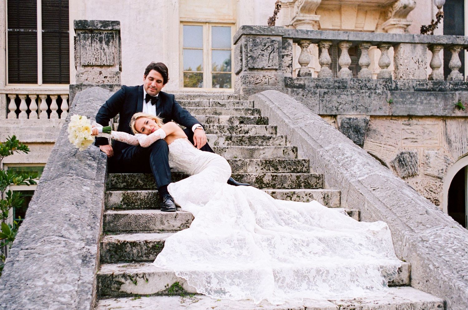 A bride in a flowing white dress poses on ornate stone steps at a historic mansion.
