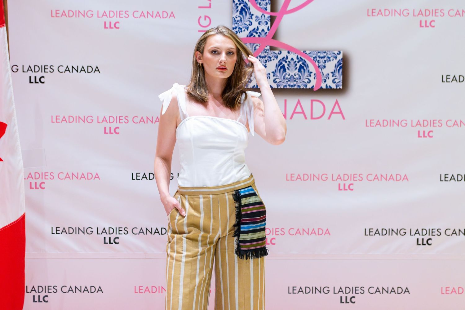 A person in a white top and flowing tan palazzo pants poses against a pink step-and-repeat backdrop with brand logos.
