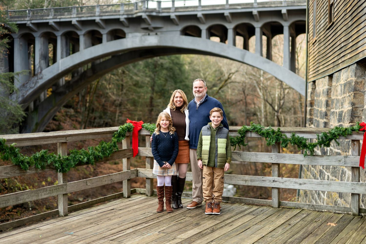 Family of four poses on decorated wooden bridge with arched stone bridge backdrop during Christmas season.