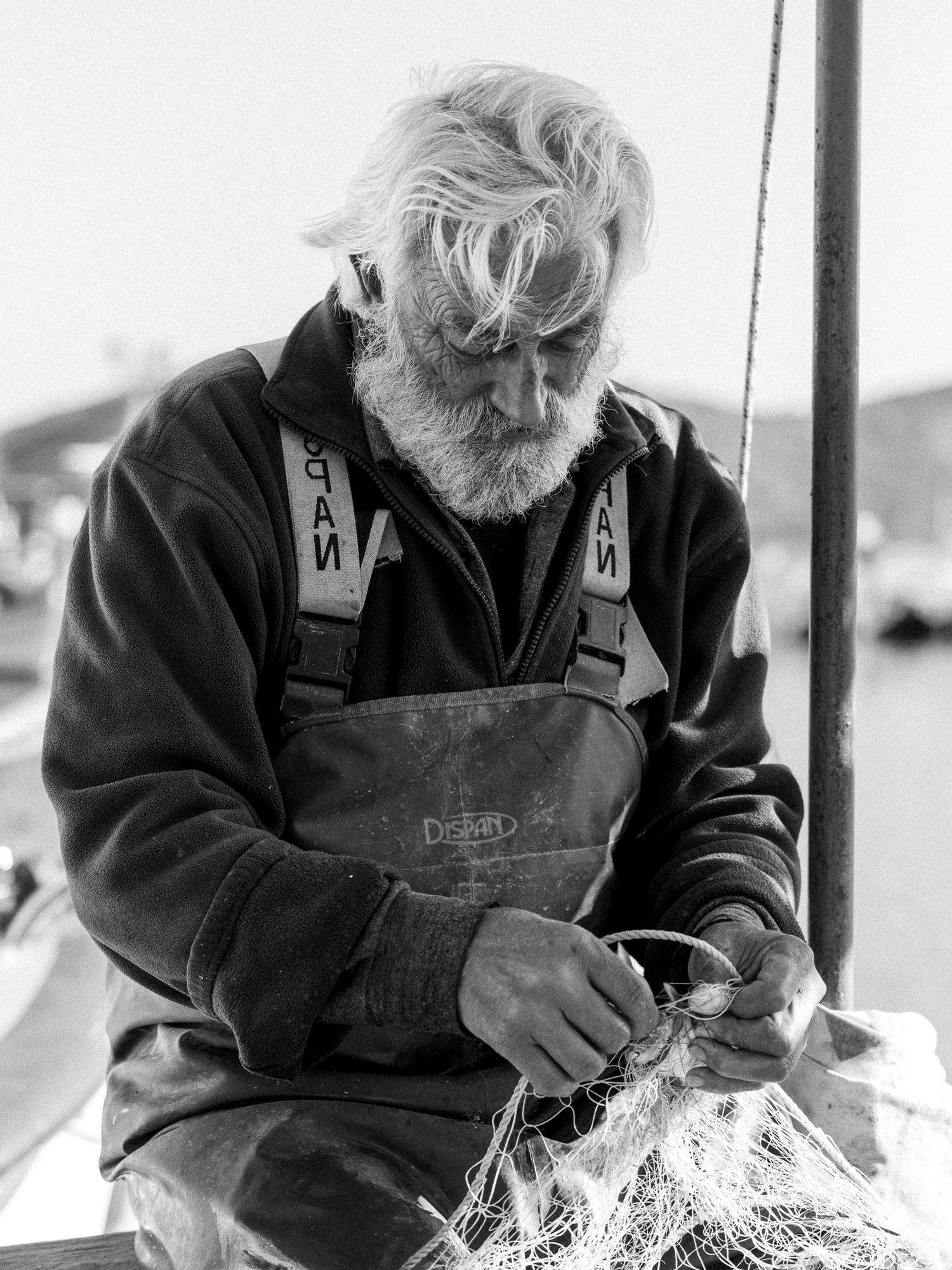 Elderly fisherman in Elounda, Crete repairing yellow fishing nets on his boat, documentary black and white portrait by the sea.