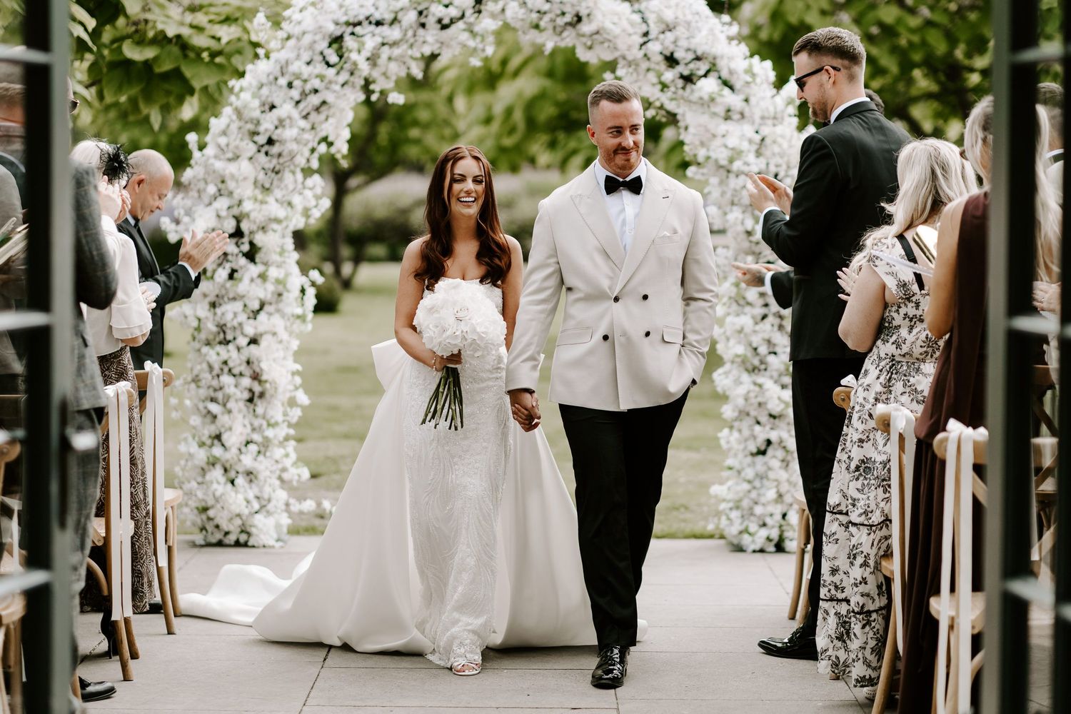 Wedding party walks down a garden path lined with elegant white cherry blossom trees and floral arrangements.