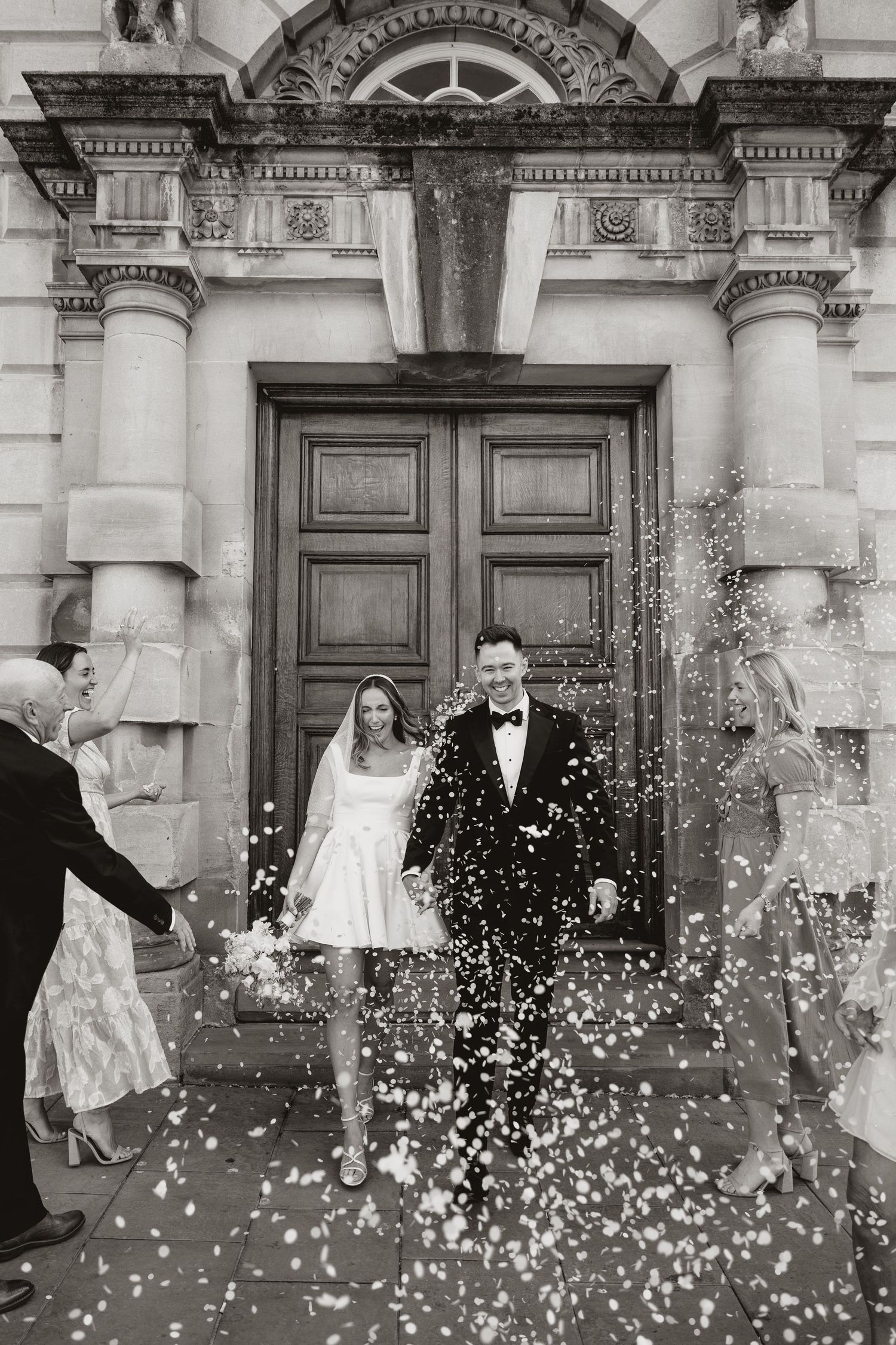 Black and white wedding photograph showing a couple in formal attire standing in a grand doorway as guests throw confetti.