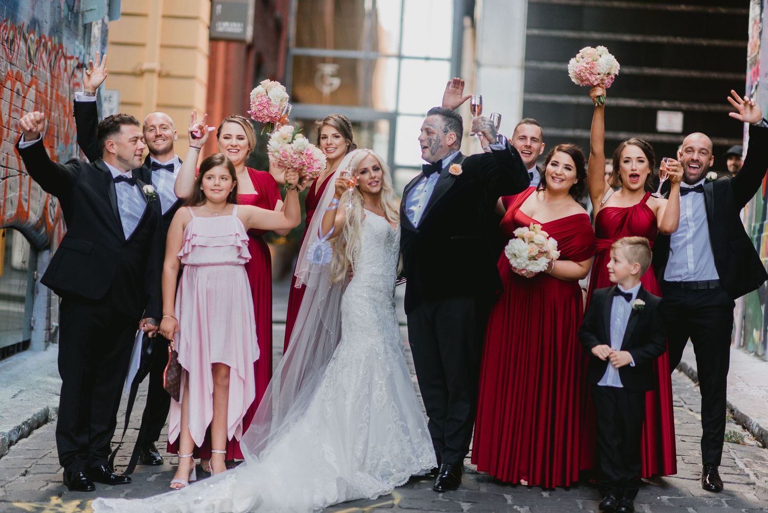 Wedding party outdoor group portrait with burgundy bridesmaid dresses and formal attire celebrating with raised arms.