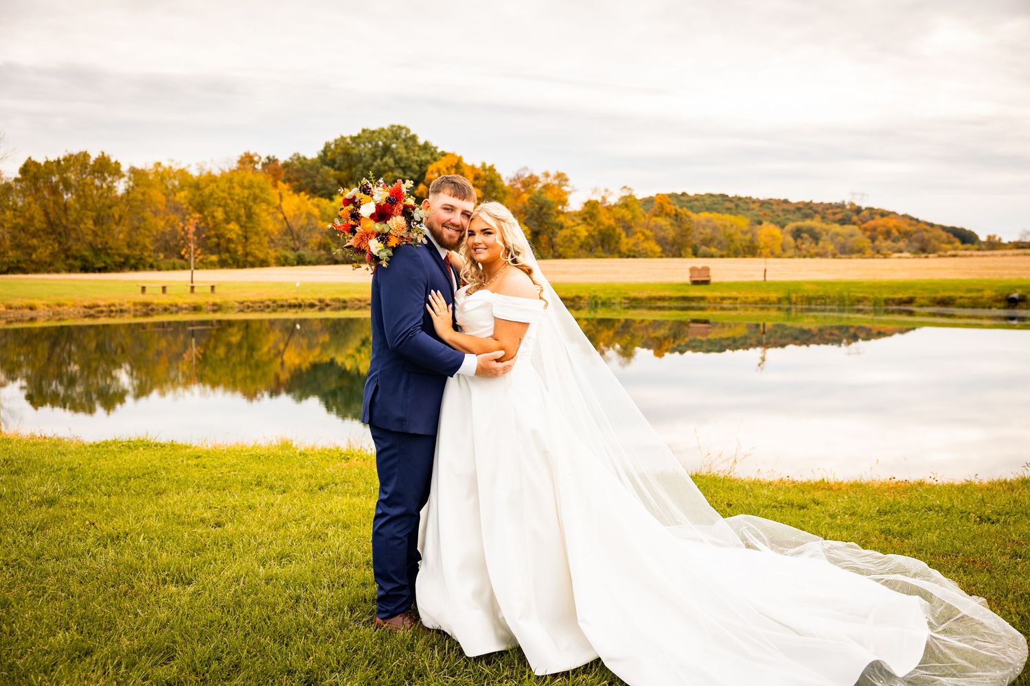 A romantic couple in wedding attire embraces near a scenic lake during golden hour at sunset.