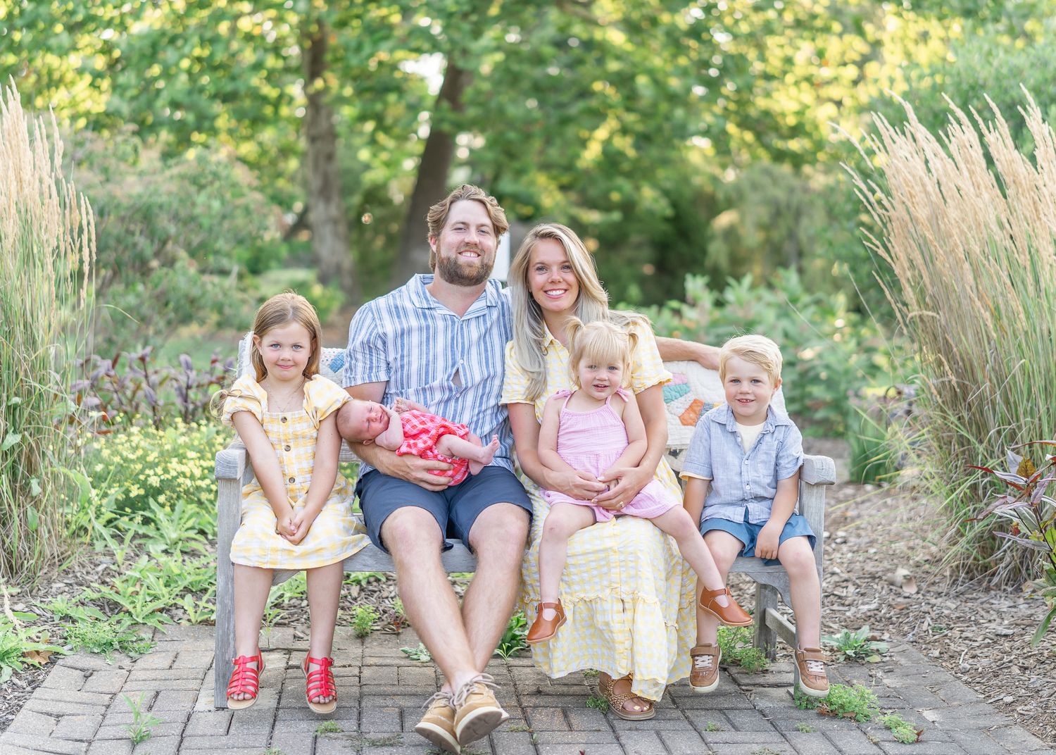 A family sits together on a wooden bench surrounded by tall ornamental grasses and greenery on a brick path.