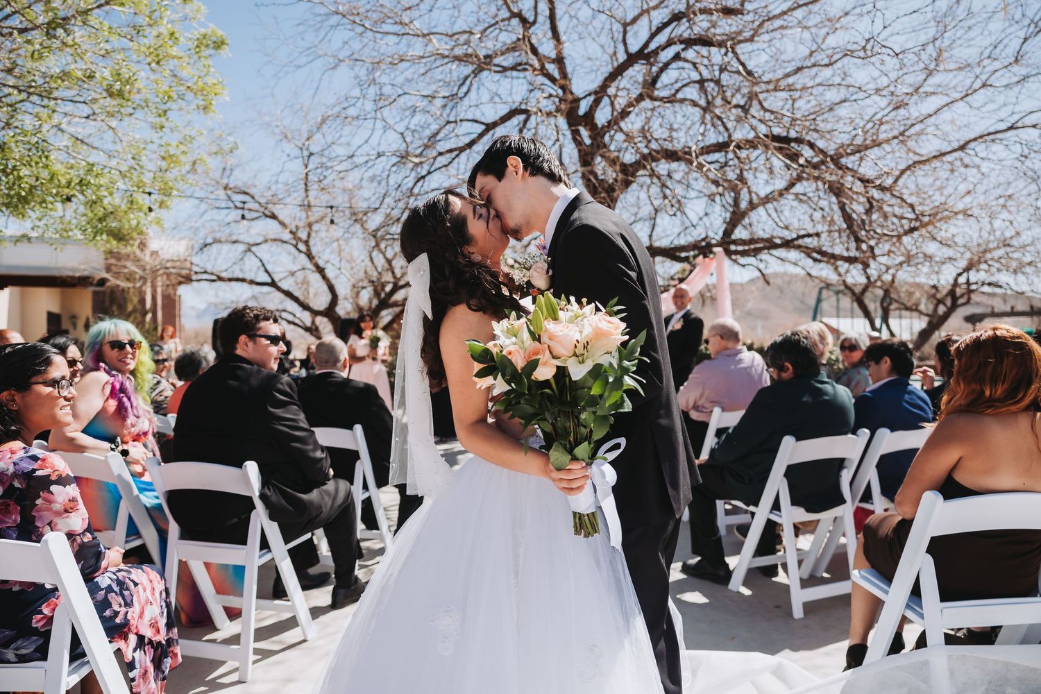 A couple shares a romantic moment during their outdoor wedding ceremony surrounded by guests in white folding chairs.