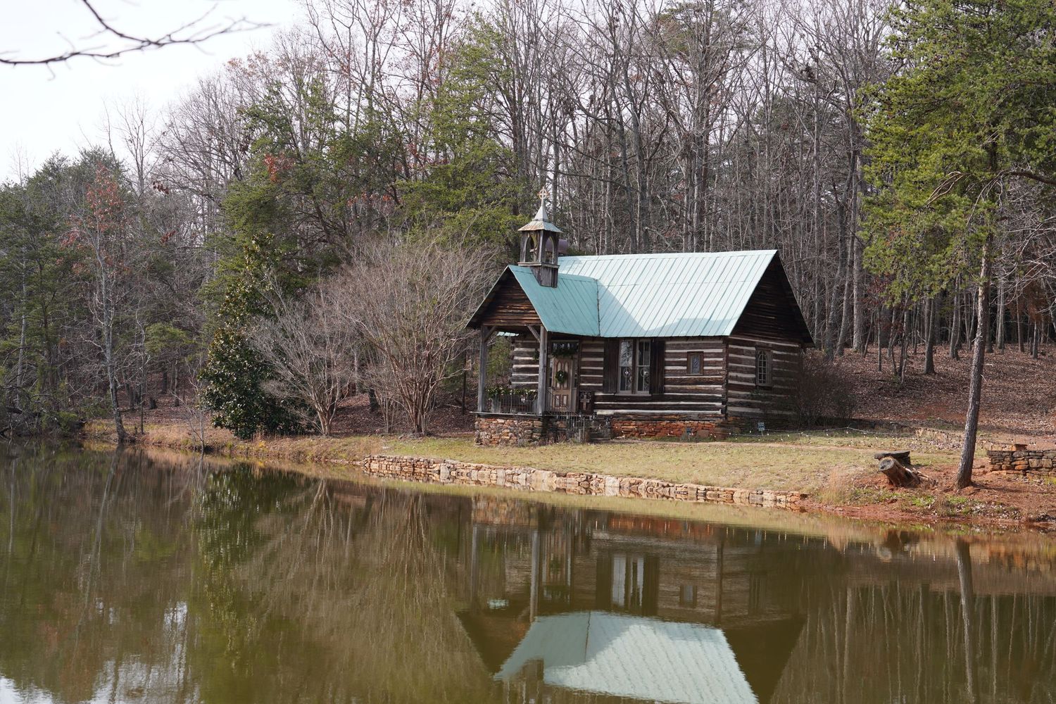 A scenic nature trail leads to a rustic log cabin nestled beside a tranquil pond in a wooded forest setting at Hart Square Village.
