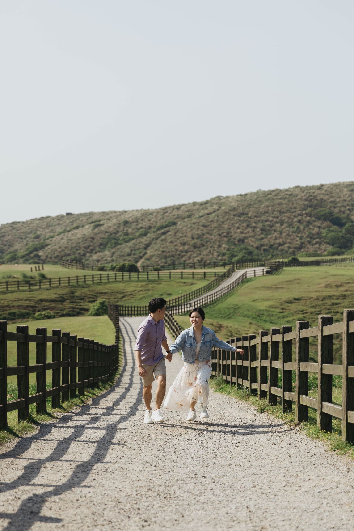Young couple holding hands running along a scenic gravel path flanked by wooden fences and green hills.