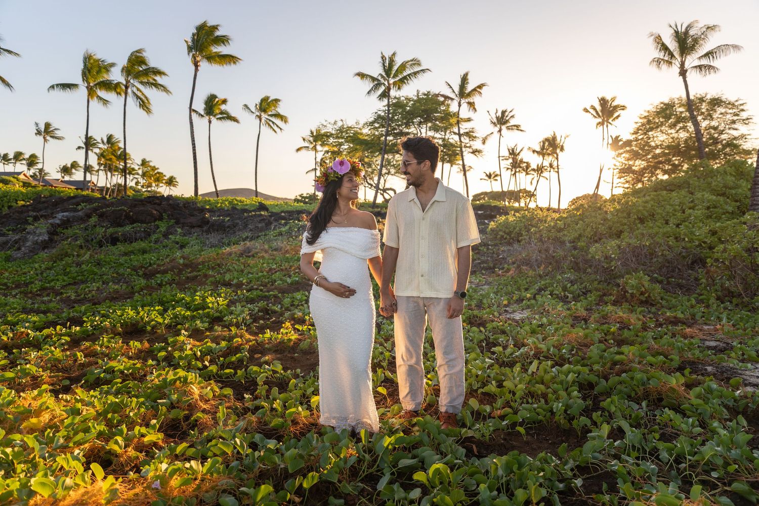 A couple stands together on a tropical hillside with palm trees and golden sunset light in Hawaii.