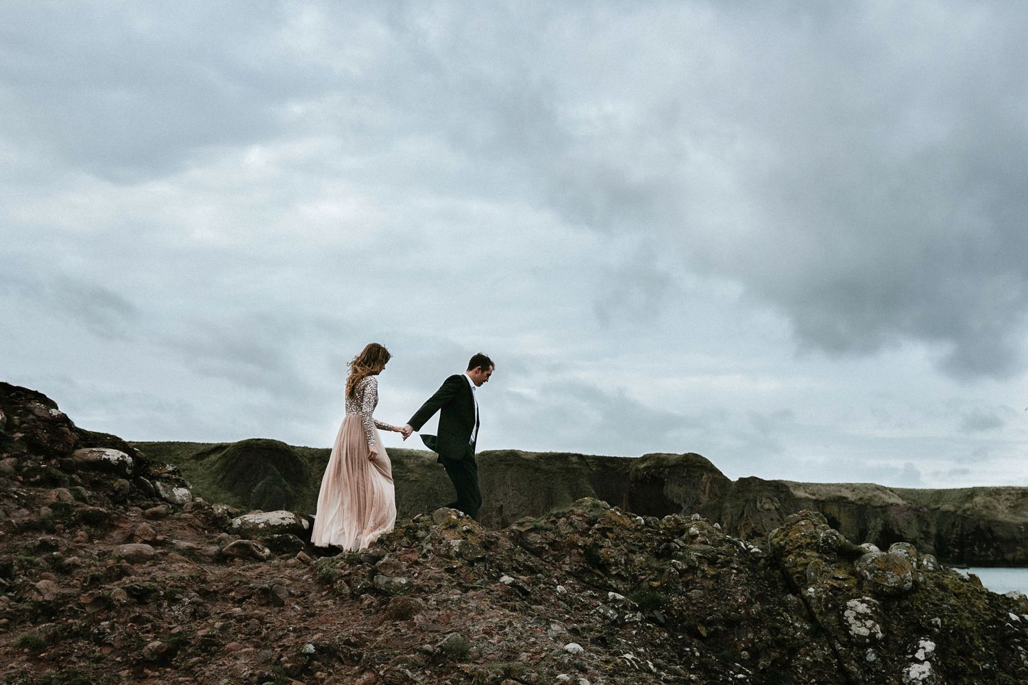 Couple holds hand as they walk across a rocky coastline during engagement photography session.