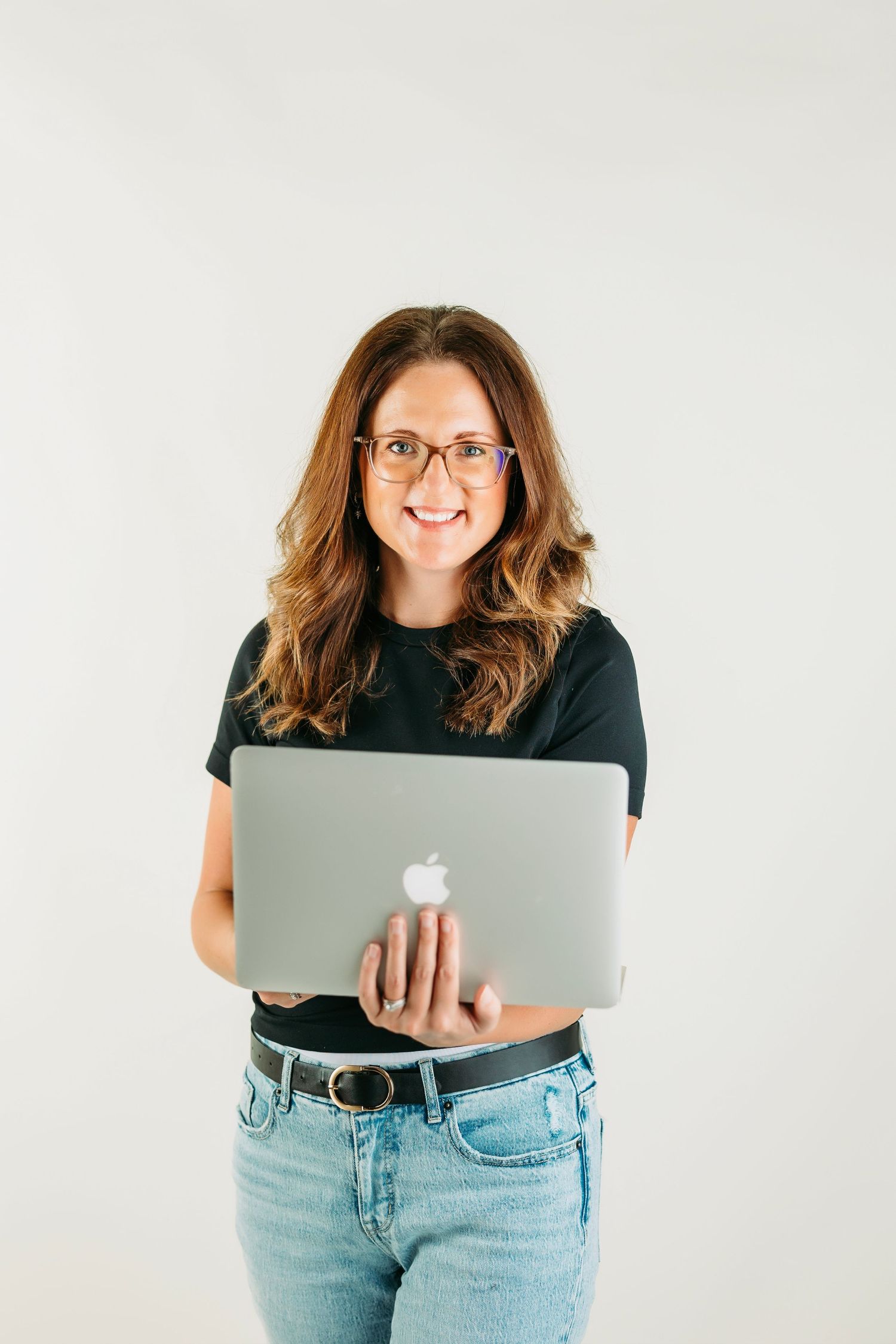 A person wearing glasses and casual attire stands against a white background while holding a MacBook laptop.