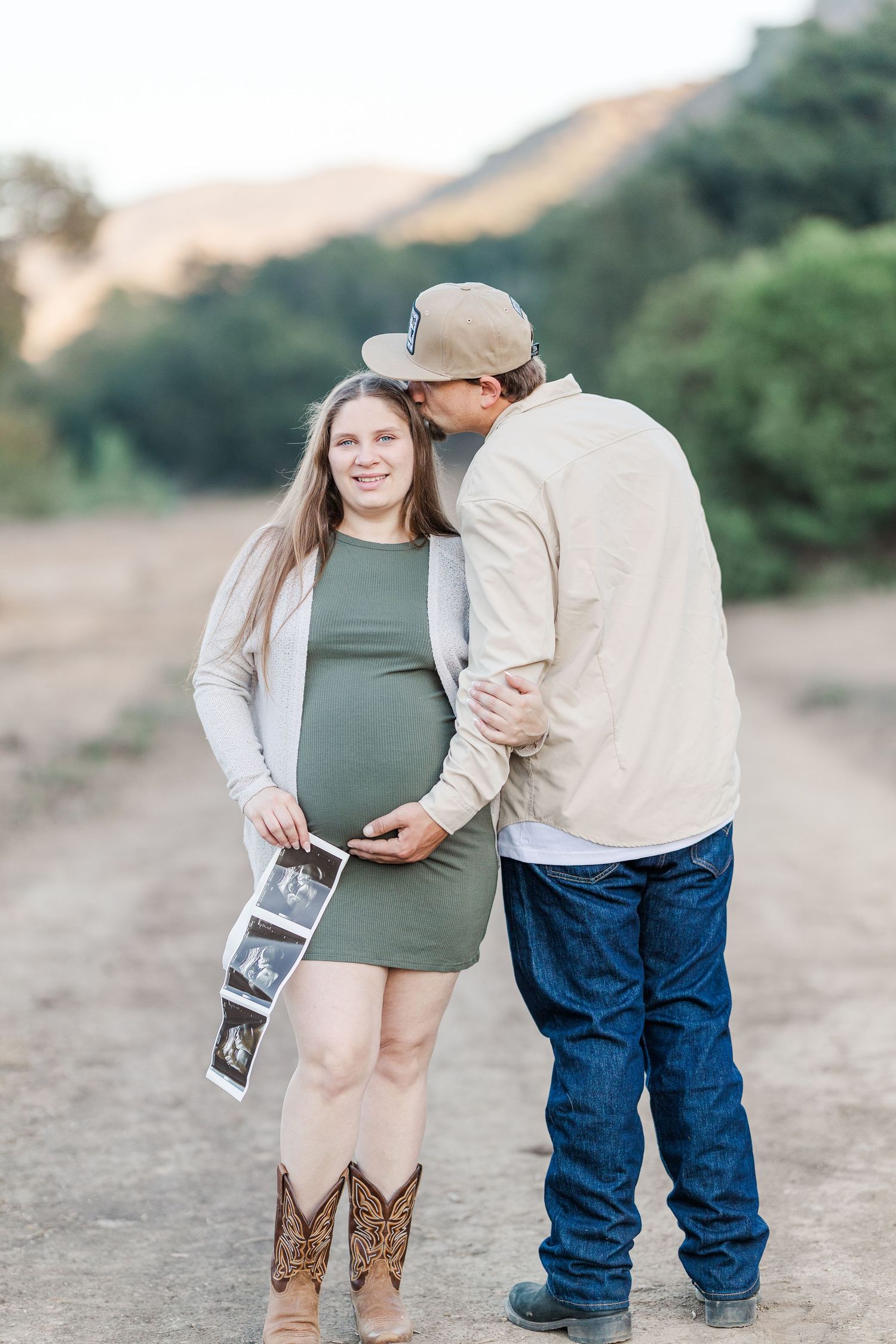 A couple embraces on a dirt path while holding ultrasound photos during a maternity photoshoot in nature.