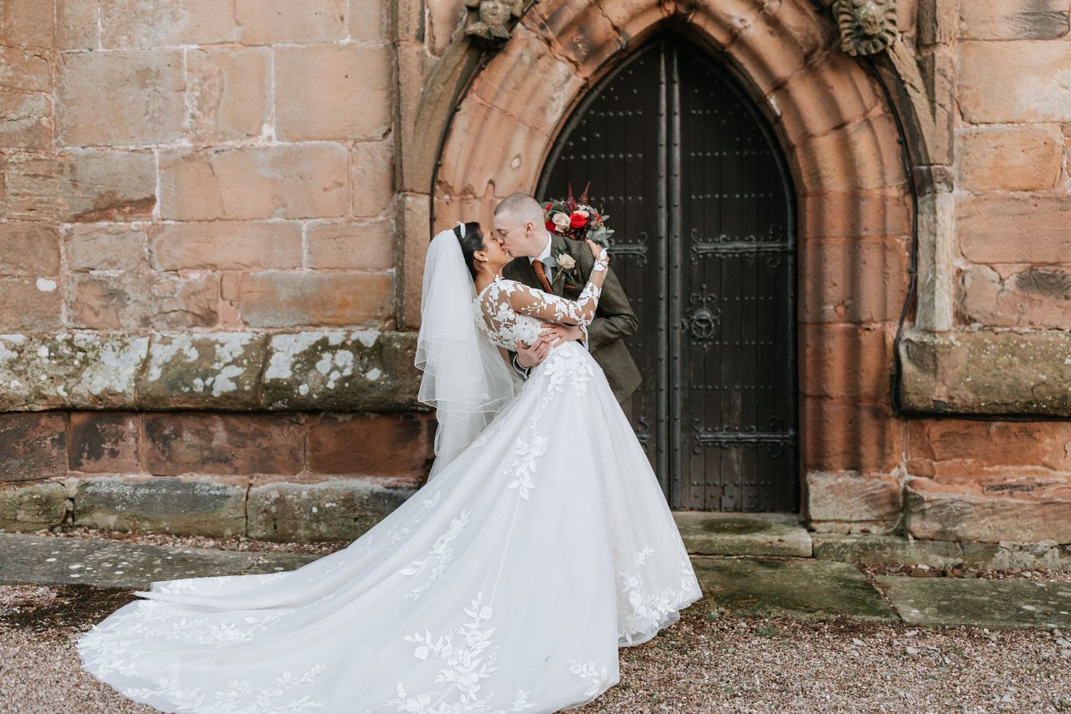A couple in wedding attire embraces in front of a rustic stone church entrance with arched doorways.