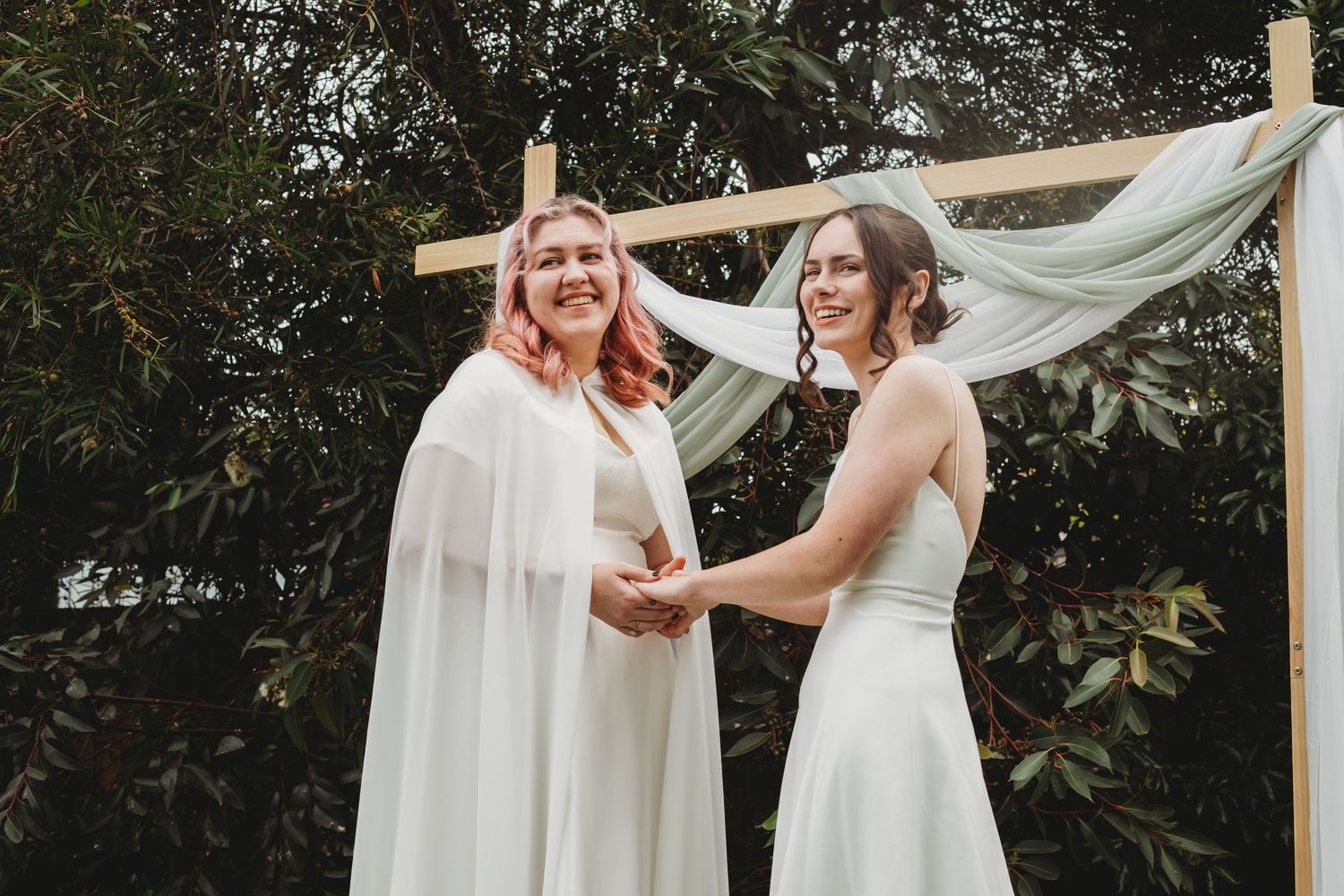 Three brides wearing white gowns gather together outdoors beneath a flowy white canopy at a wedding ceremony.
