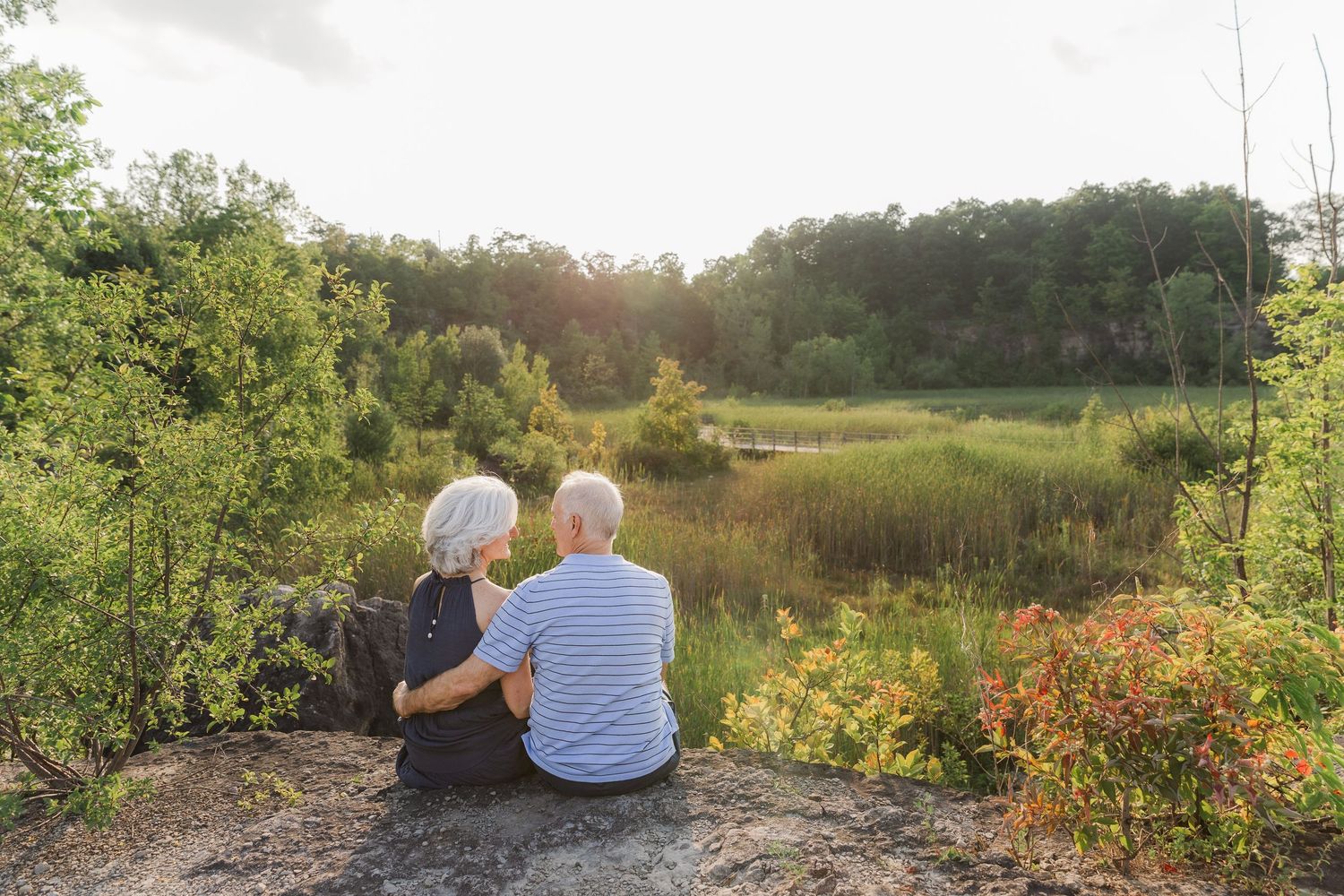 Two people sit close together watching a sunset over a scenic natural landscape.