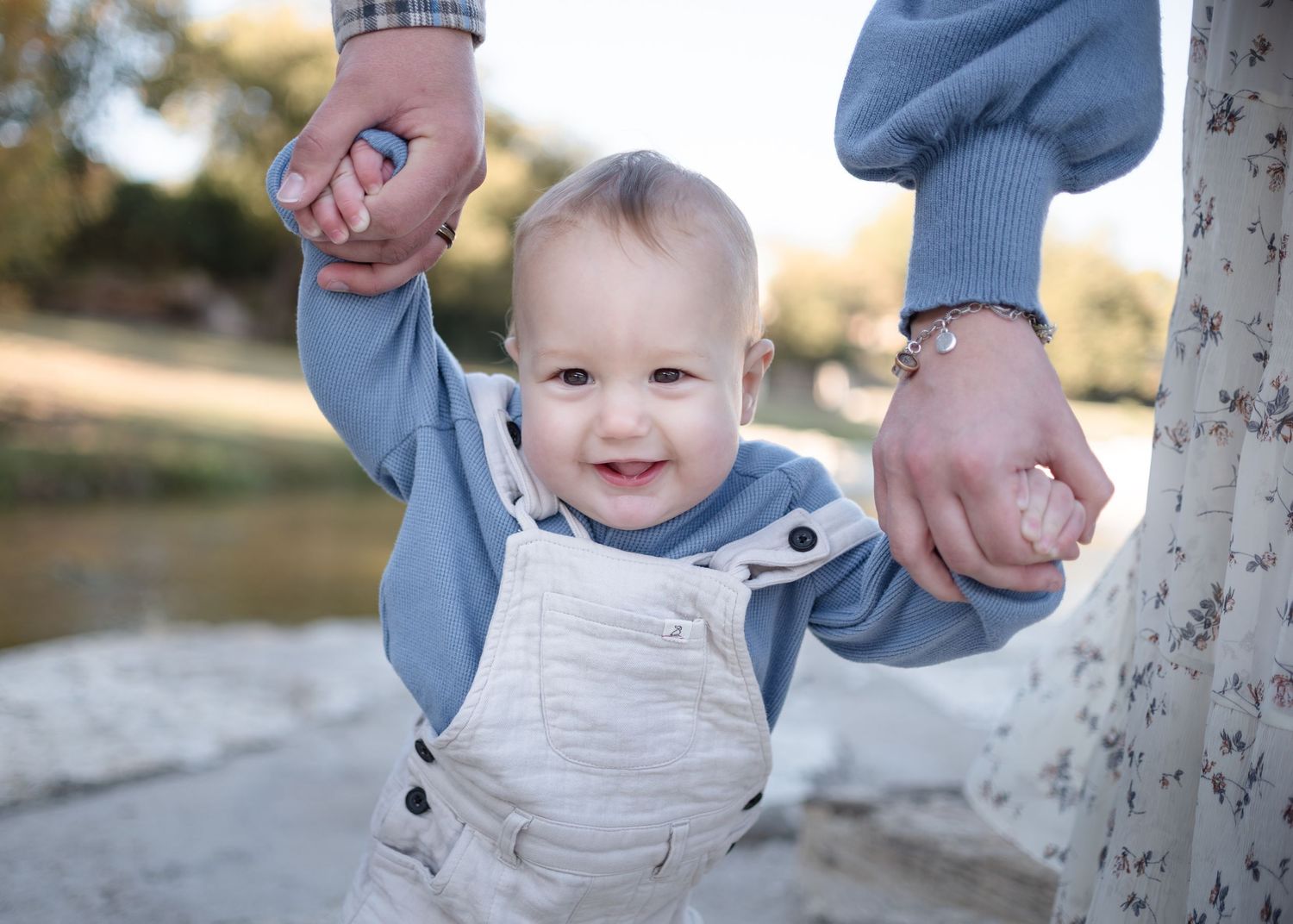 Parents holding baby’s hands as he walks during outdoor family session at Nolan Creek in Belton Texas