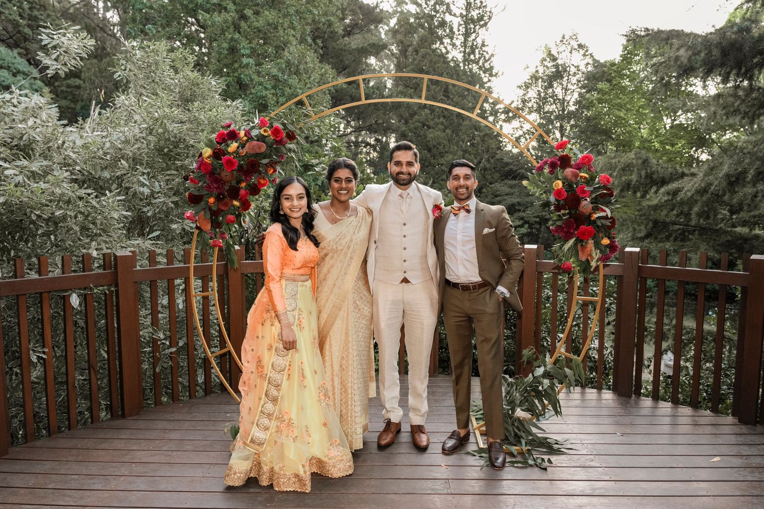 Wedding party stands under floral arch on wooden deck decorated with red roses and greenery.