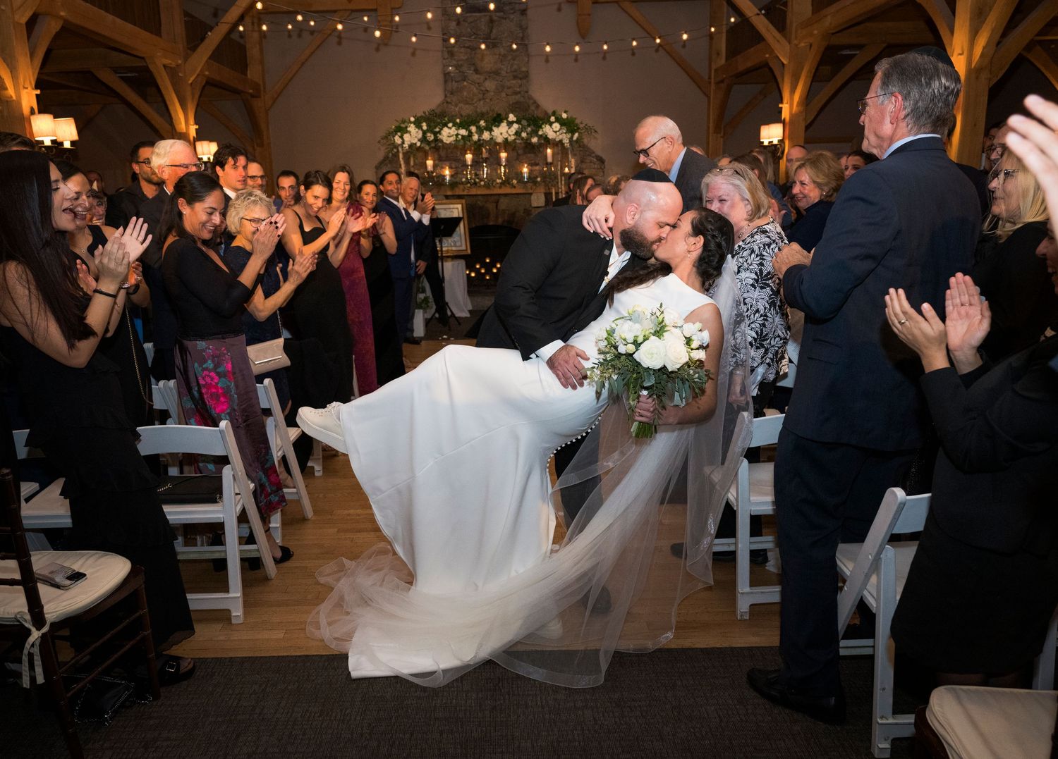 Wedding guests dance and celebrate on the dance floor under twinkling lights at a rustic barn reception venue.