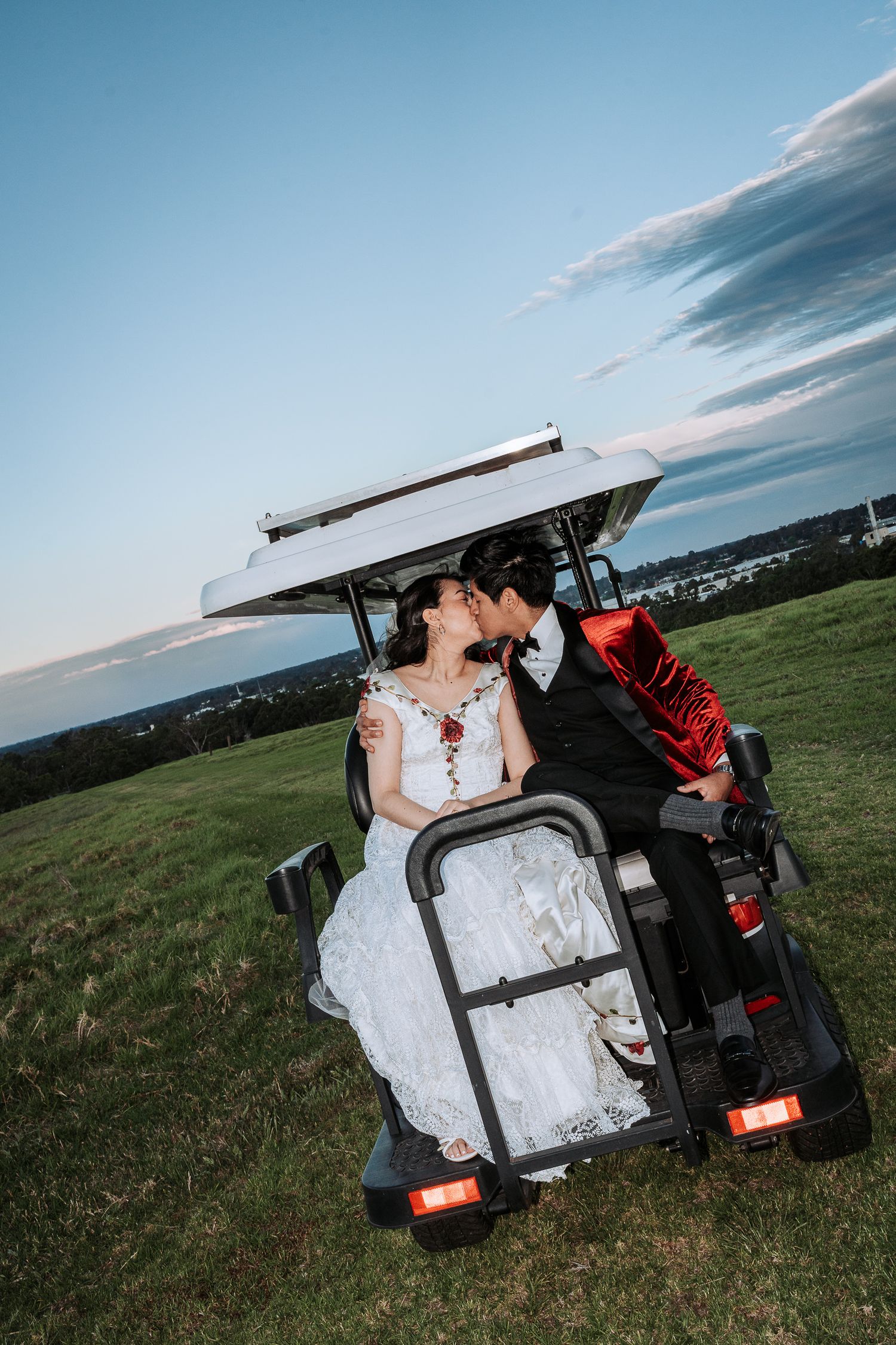 A couple in wedding attire share a romantic moment in a golf cart on a hilltop overlooking scenic views at sunset.
