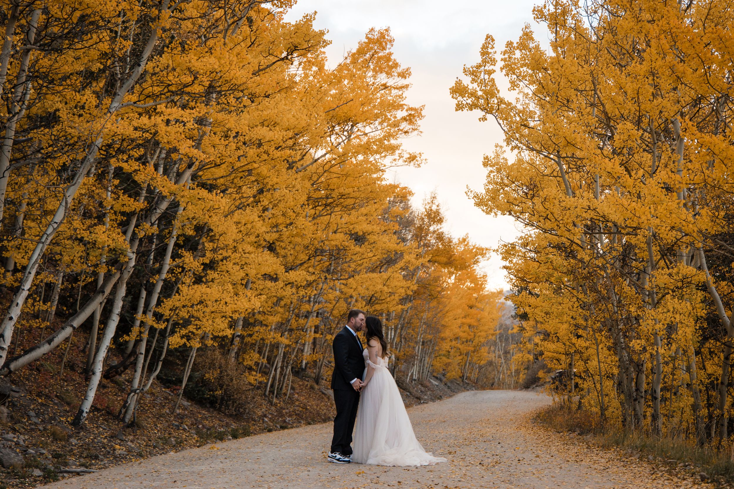 A couple stands on a dirt road surrounded by golden aspen trees during autumn sunset.
