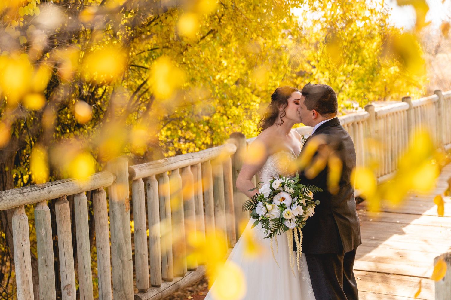 Romantic couple embracing under vibrant yellow autumn foliage with wooden fence in background.