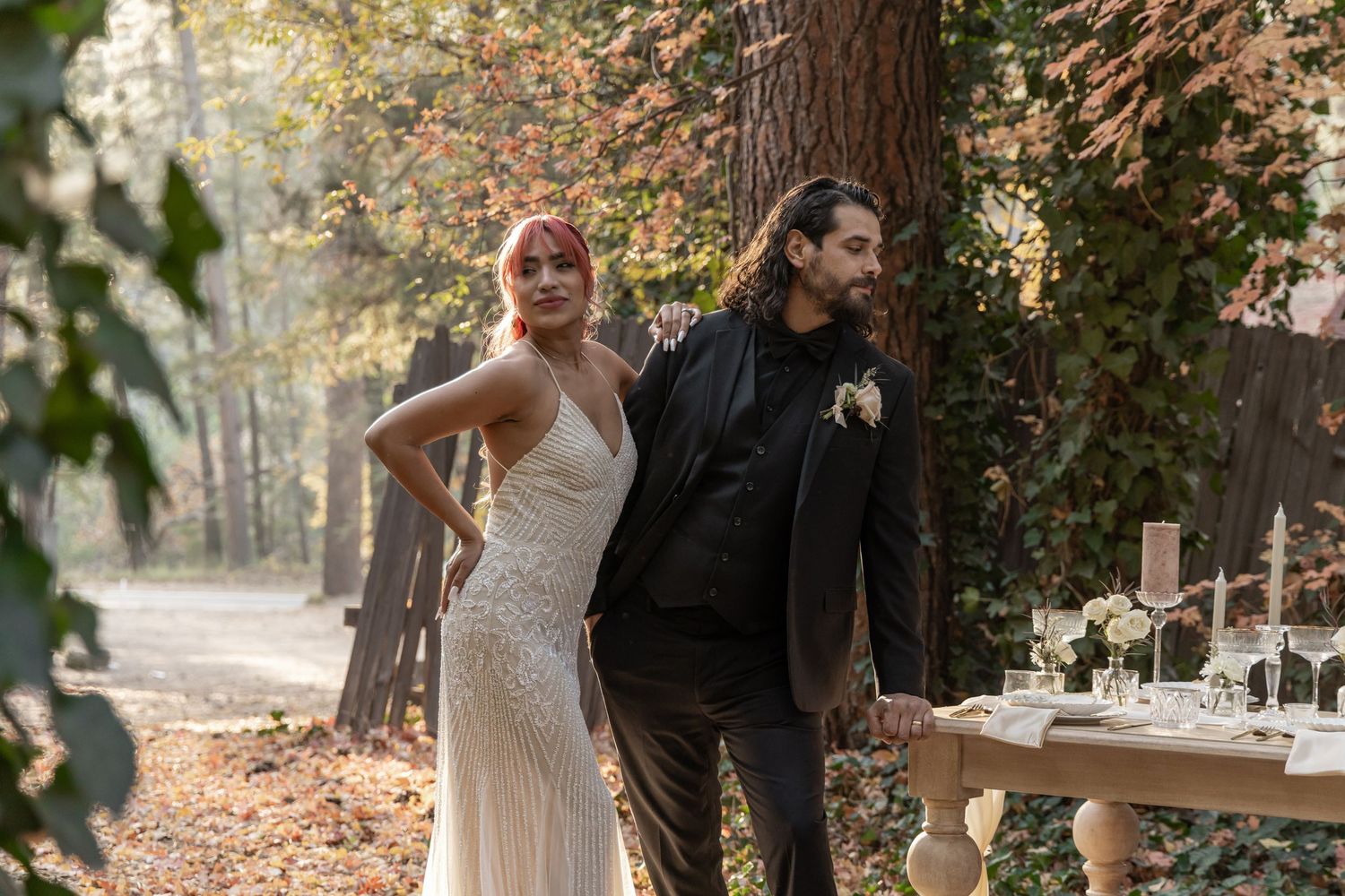 A rustic outdoor wedding reception setup with long tables and white tablecloths under tall trees in an autumn forest.