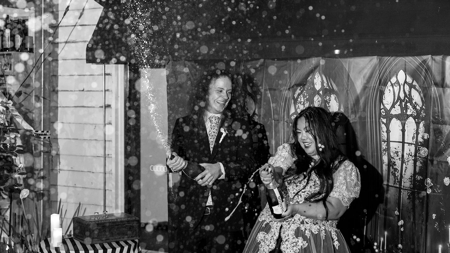 A black and white photograph shows a Bride and Groom spraying champagne during their wedding speech during their Halloween wedding.