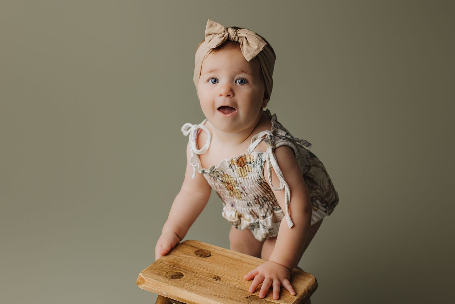 Baby in floral dress and bow crawls and plays on wooden floor in natural light studio setting.