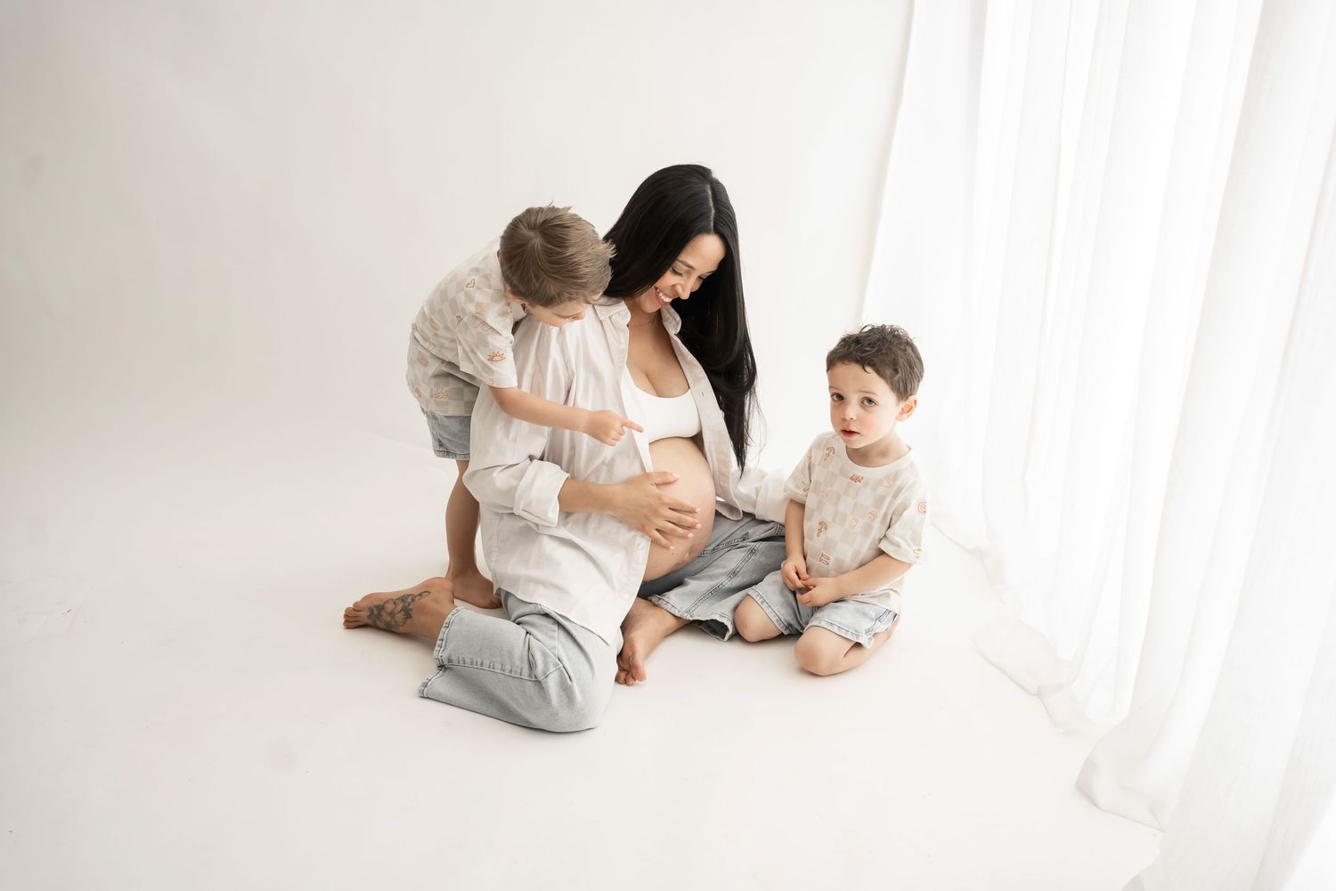 Pregnant woman in white blouse sits sideways on sleek black barstool against minimalist white background.