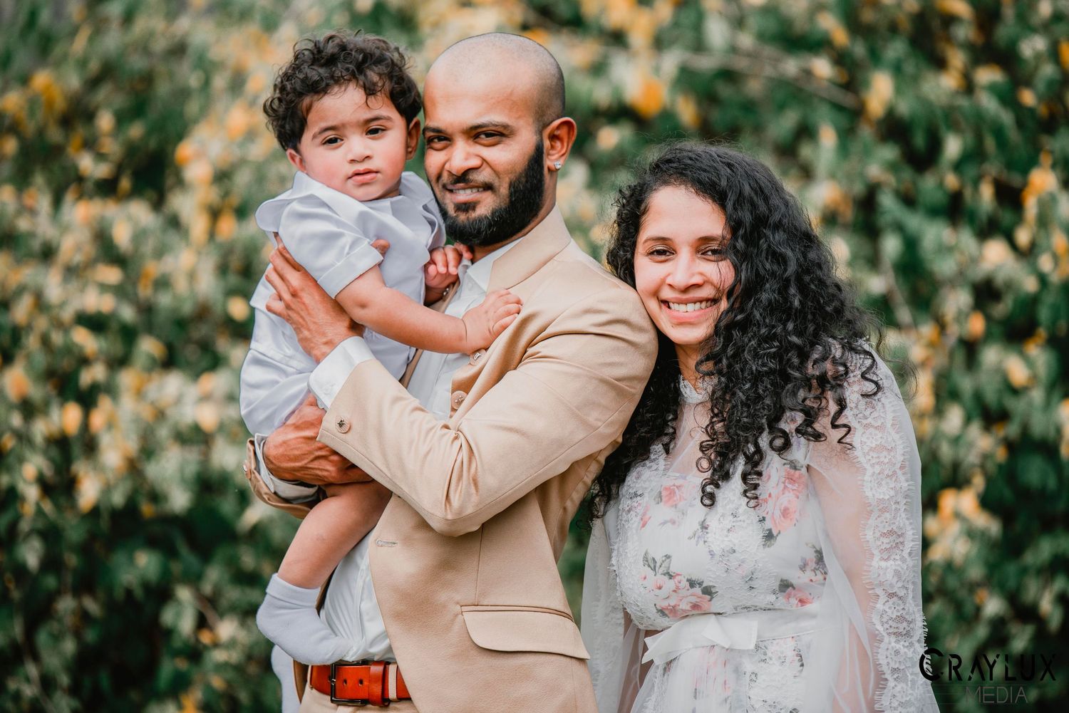 A family poses together outdoors in front of green foliage during a sunny autumn day.