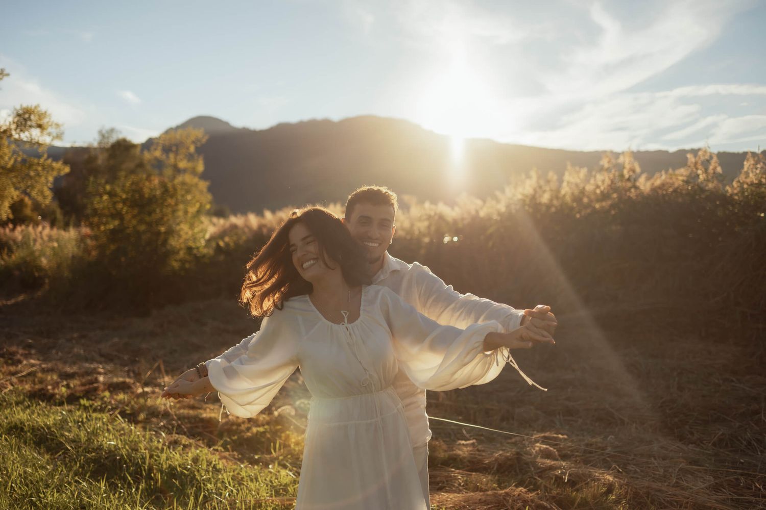 A joyful couple embraces in golden sunset light, the woman in a white dress with arms outstretched in a field.