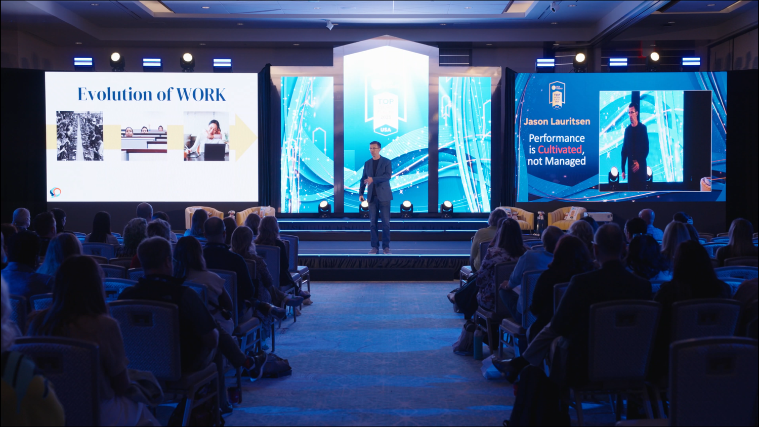 Conference attendees seated in a dark auditorium with multiple presentation screens displaying business content.