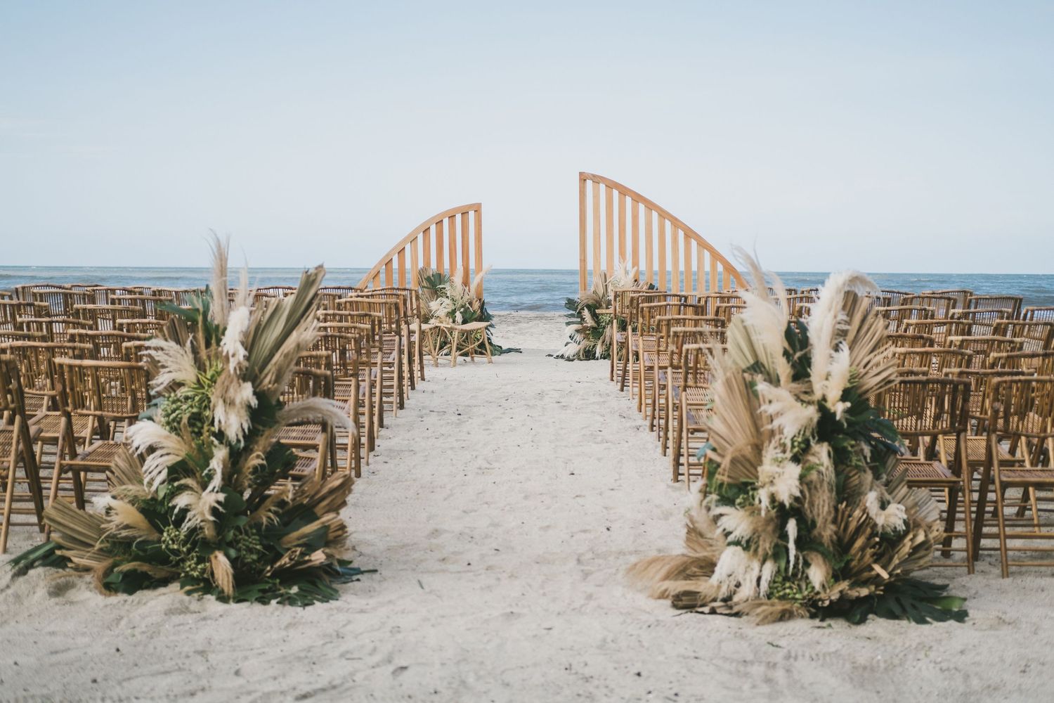 A beachfront wedding ceremony setup with wooden arch and chairs decorated with pampas grass and dried floral arrangements.
