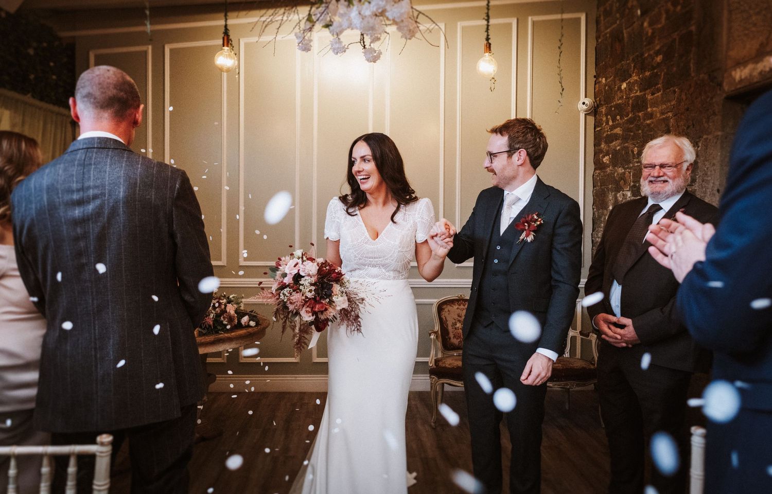 Wedding guests celebrate with bubbles as newlyweds exit through a grand hallway with elegant wall paneling.