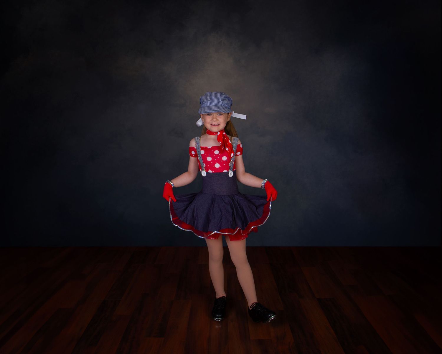 A dancer in a polka dot dress and blue hat performs against a dark backdrop on a wooden floor.