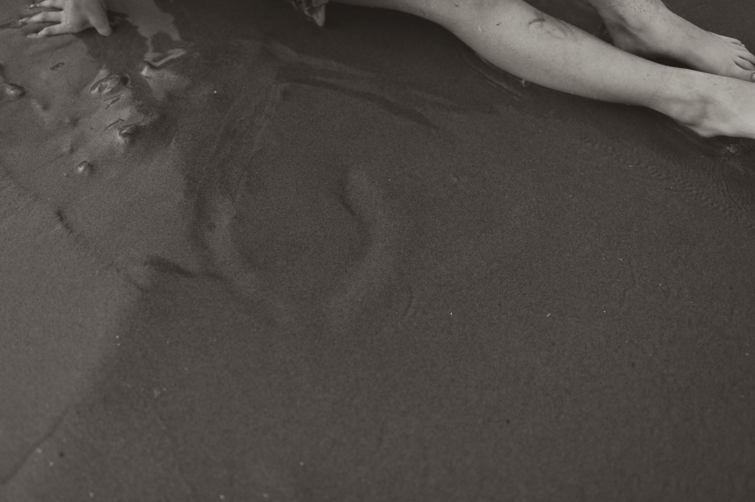 Black and white photo of bare feet and hands touching wet sandy beach shore with water ripples.