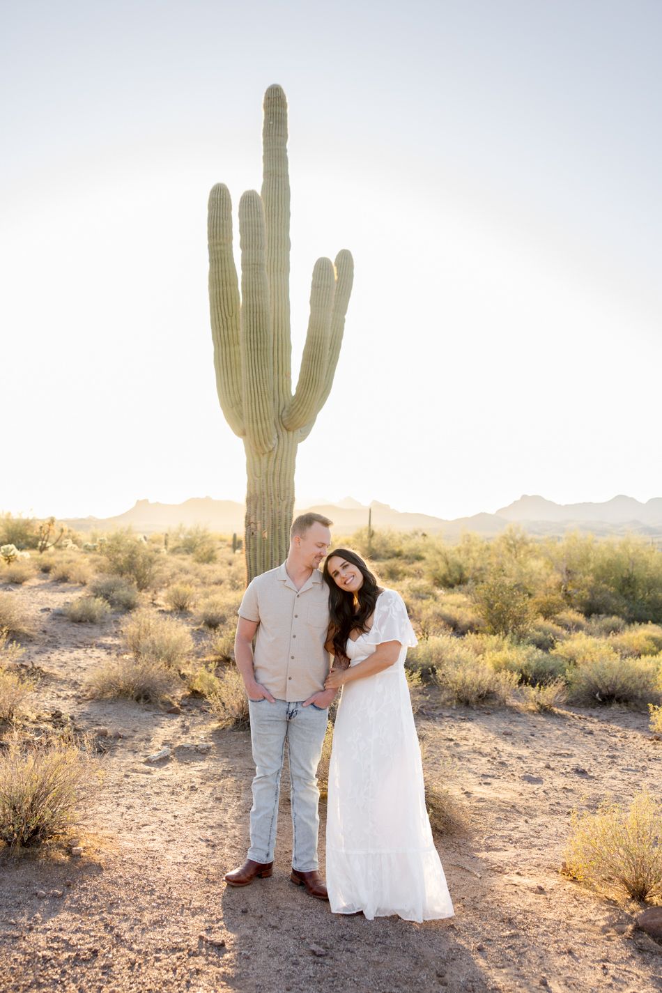 A couple stands together near a tall saguaro cactus in the Arizona desert during golden hour.