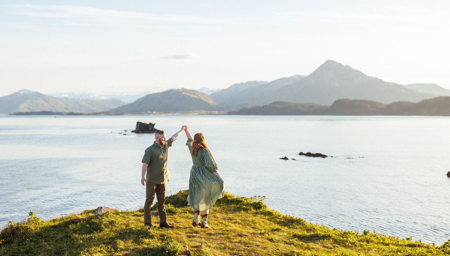 A couple holds hands while standing on a wooden dock overlooking a misty lake with evergreen forests in the background.