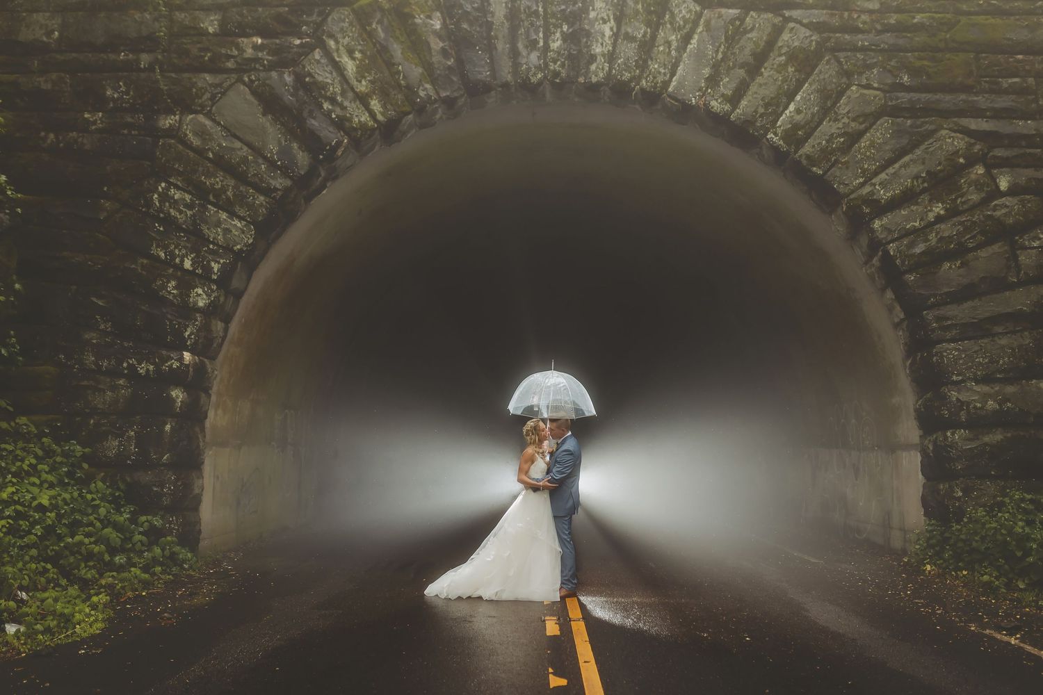 Wedding couple under stone bridge archway holding white umbrella.