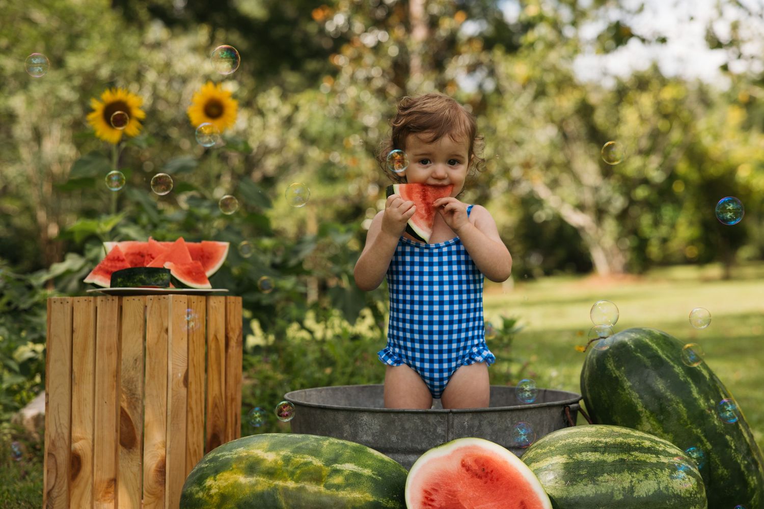 A small child in a checkered swimsuit reaches for bubbles in a sunny garden with watermelon and sunflowers nearby.