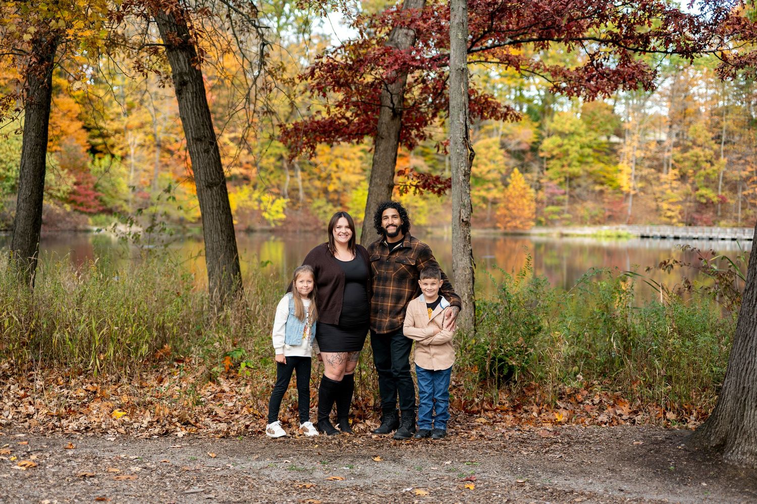 Family of four poses by a colorful autumn lake, parents with two young children among vibrant fall foliage.