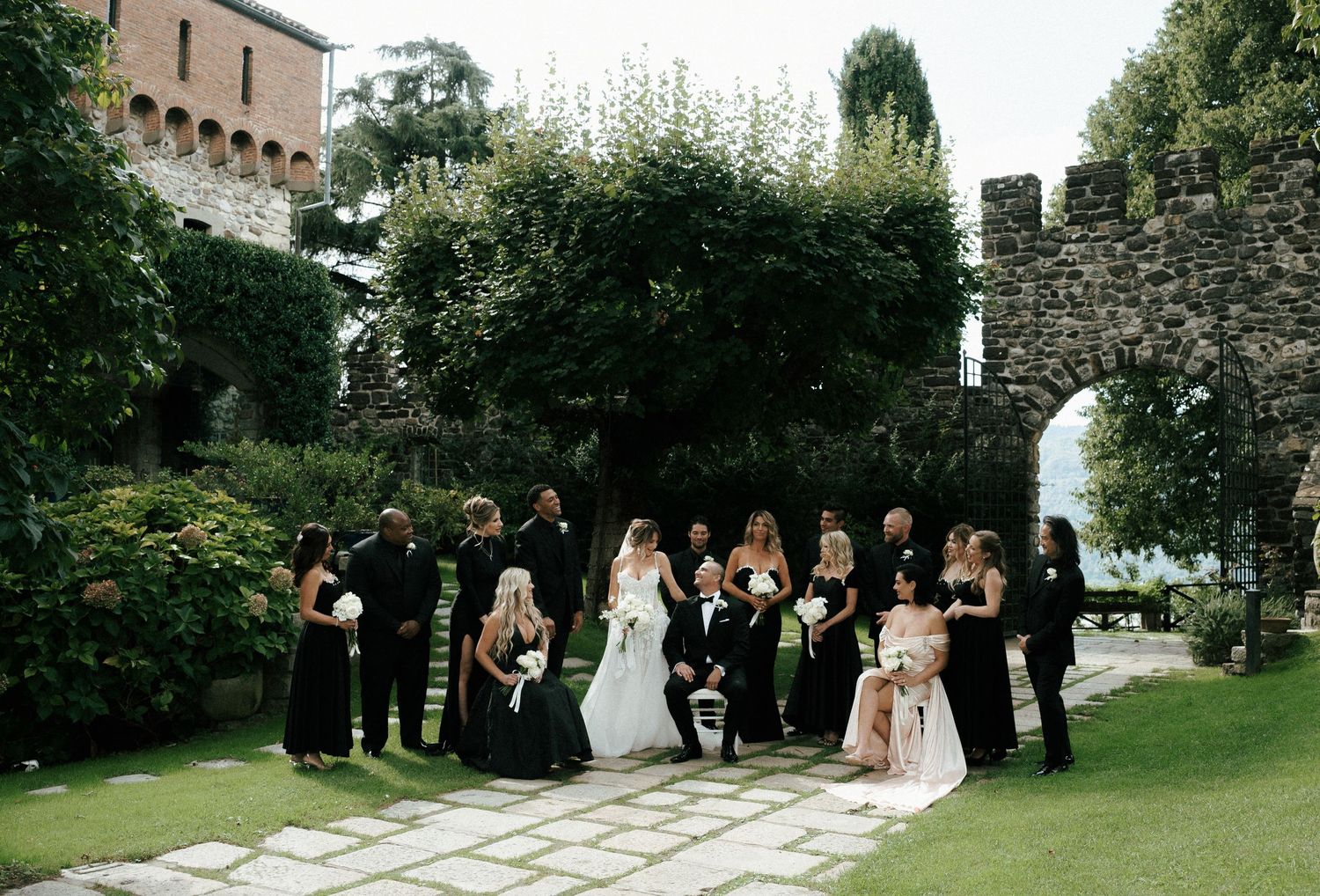 Wedding party walks in procession through lush garden pathway at historic stone castle venue.