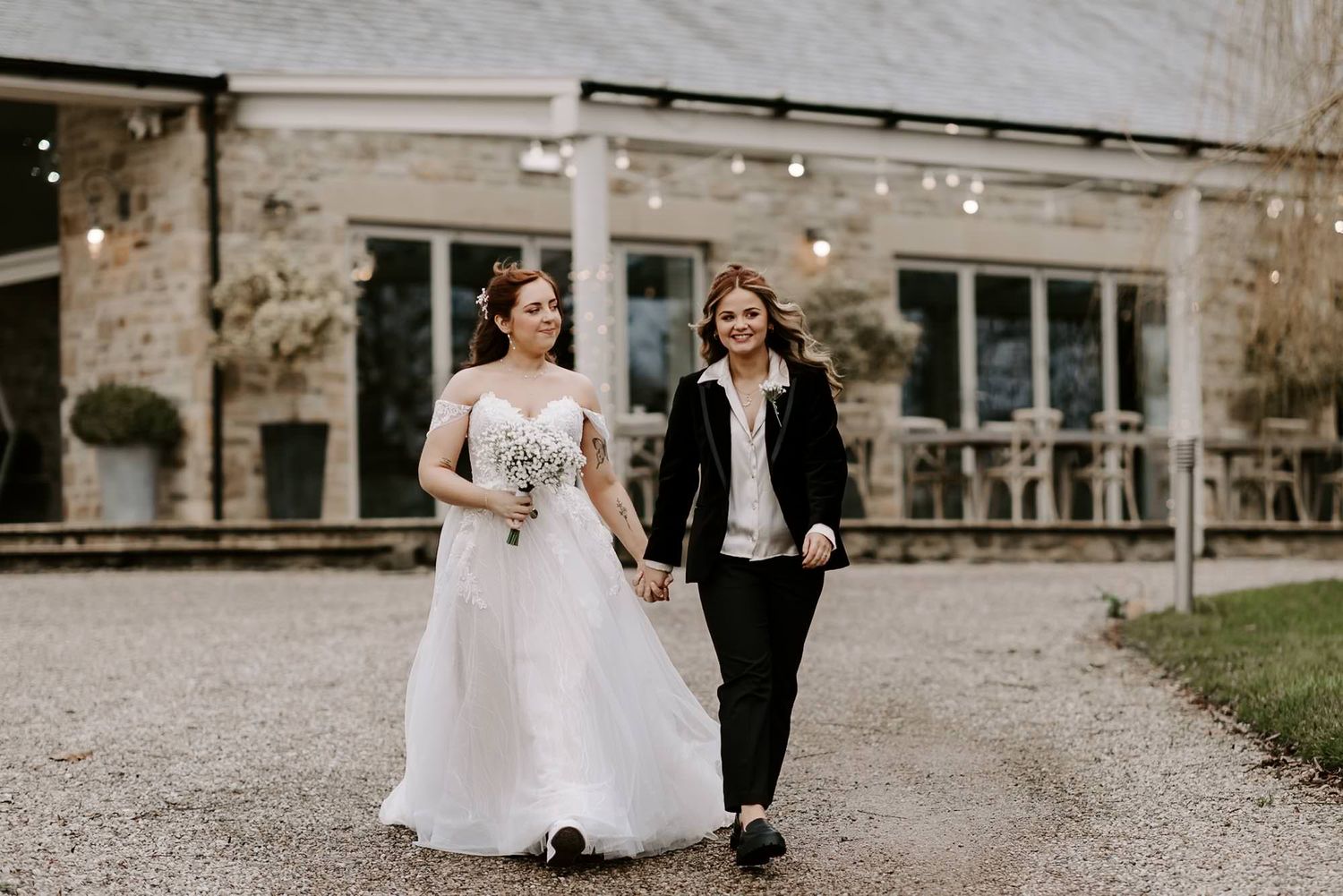 Two people walking together outdoors, one wearing a white wedding dress and holding a bouquet, the other in a black suit.