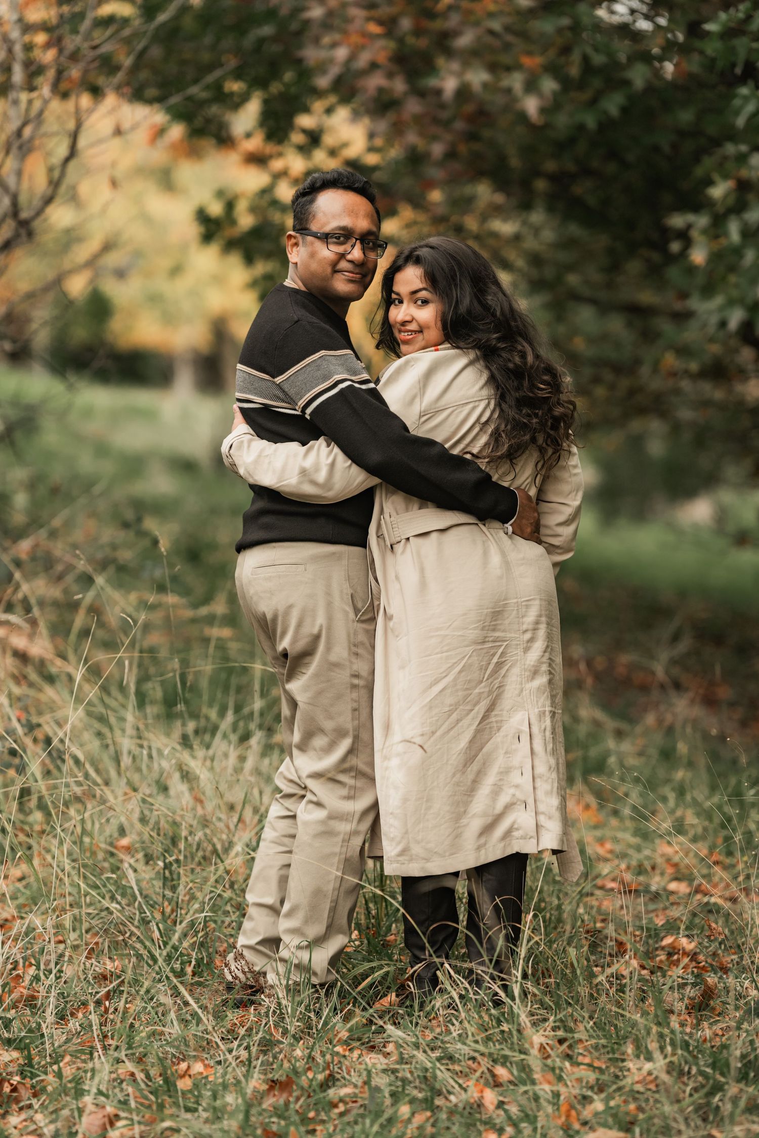 A couple embraces while standing in a field of tall grass during autumn with warm earth tones in the background.