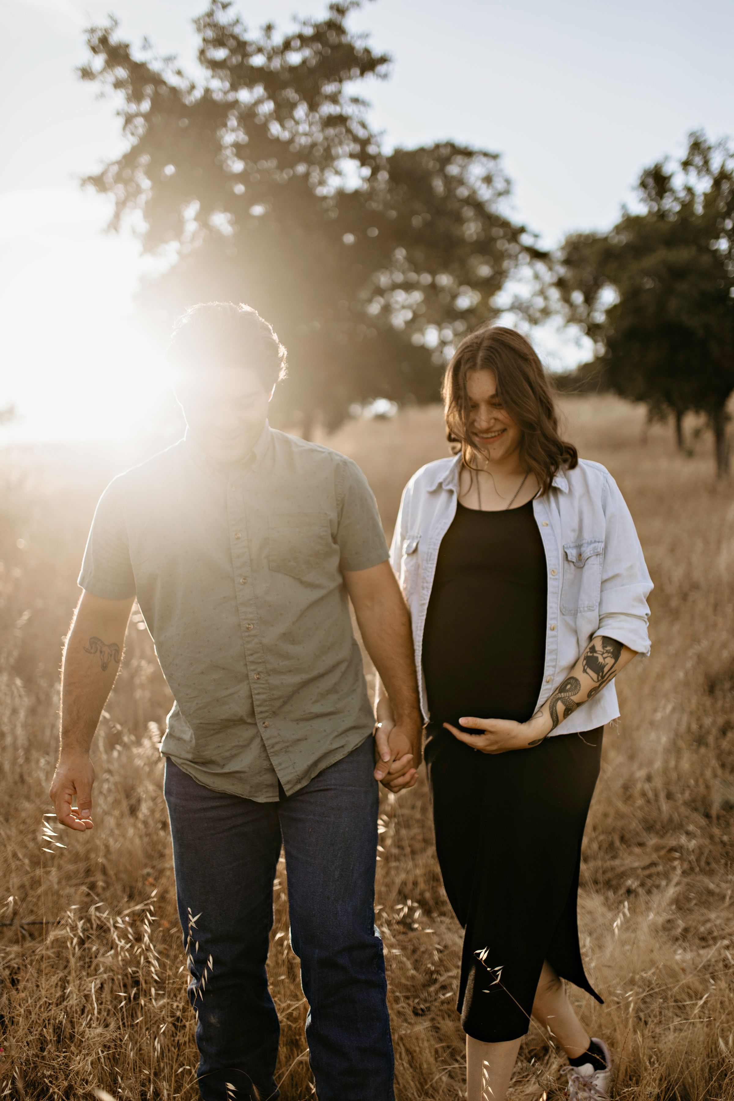 A couple in cameron Park holding hands walks through a sunlit field during golden hour as the woman gently touches her pregnant belly.