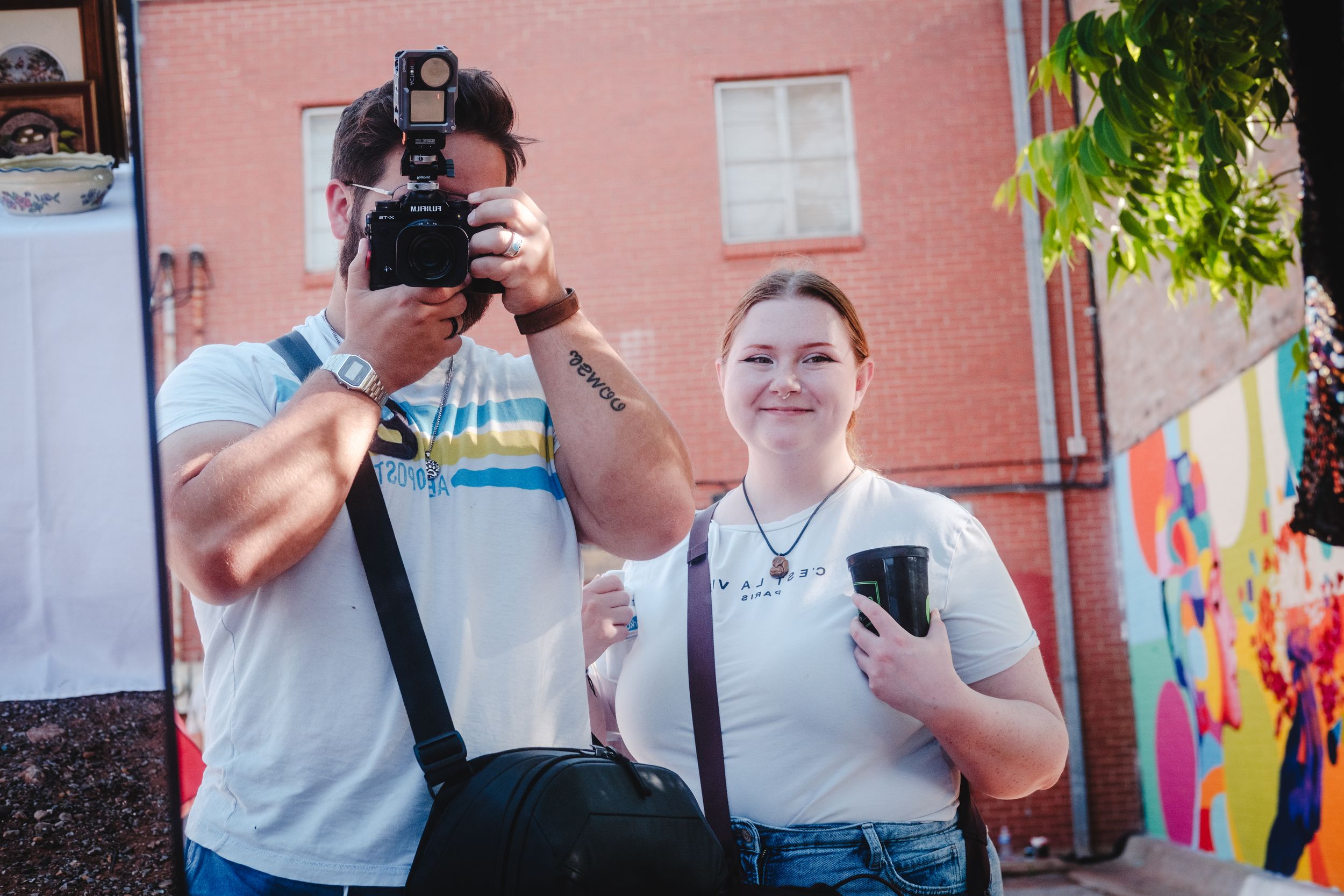 Two people stand outdoors near a pink wall, one holding a professional camera and another holding a phone.