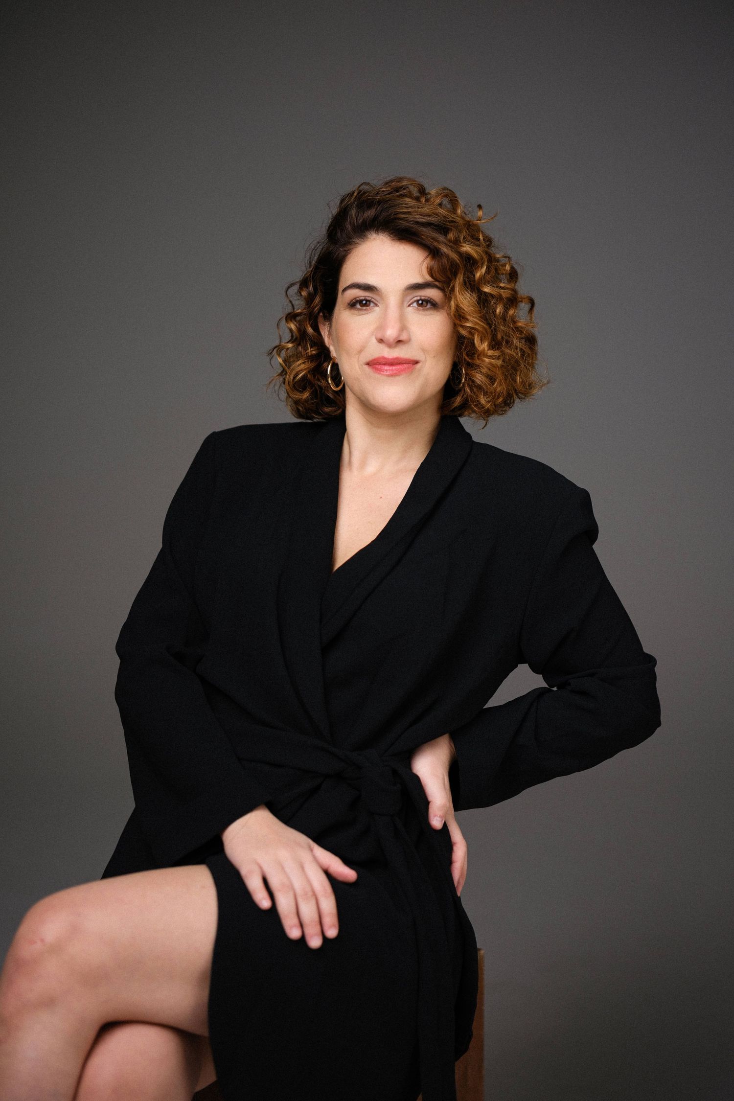 Professional studio portrait of a model in a black wrap coat with curly brown hair against a dark background.