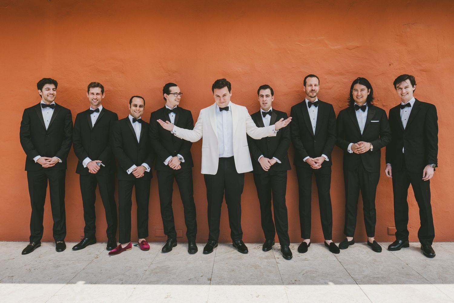 A group of nine groomsmen wearing formal black and white tuxedos stand in a line against an orange wall.