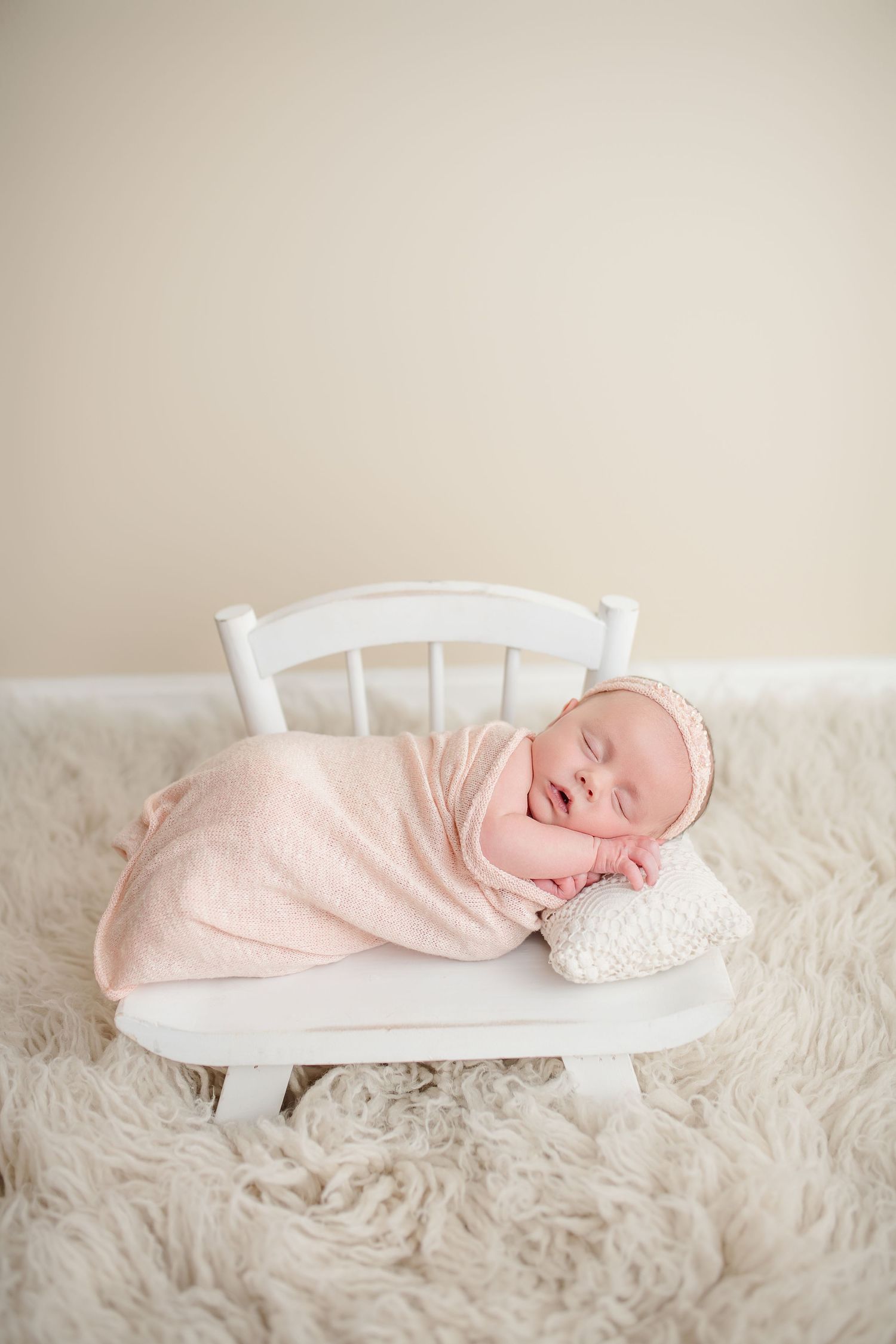 Sleeping newborn wrapped in soft pink blanket lies peacefully on small white wooden chair with plush furry backdrop.