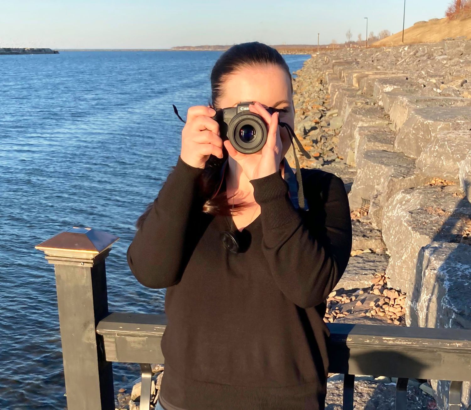 A woman stood next to a body of water, she is wearing a black sweater, her hair tied back, she has a camera up to her face